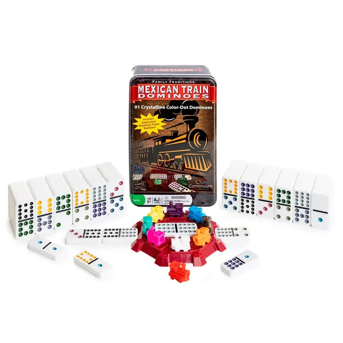 A boxed set of Mexican Train Dominoes displayed on a white background, with the metal tin box standing upright in the center and multiple white domino tiles arranged around it. The set includes colorful train-themed dominoes and a pile of small colored train pieces.