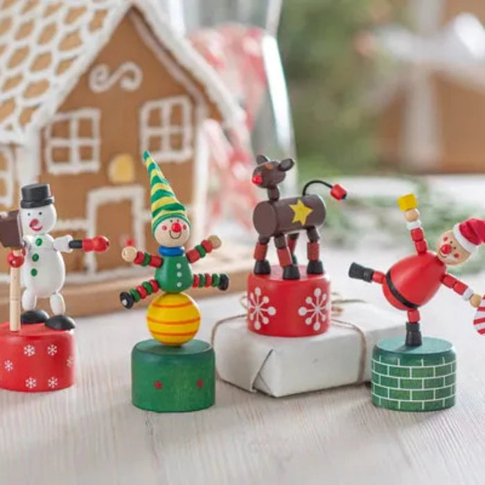 Four colorful Christmas-themed wooden figurines displayed on small painted bases in front of a gingerbread house decoration on a light tabletop.