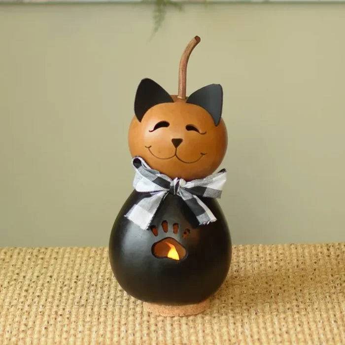 A decorative cat-shaped gourd candle holder with a black body, orange head, black ears, a black-and-white checkered bow, and a small warm light visible through a paw-print cutout, set on a woven surface against a soft beige background.