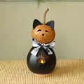 A decorative cat-shaped gourd candle holder with a black body, orange head, black ears, a black-and-white checkered bow, and a small warm light visible through a paw-print cutout, set on a woven surface against a soft beige background.