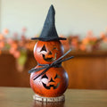 A decorative two-tiered gourd Halloween lantern with carved jack-o'-lantern faces, a black pointed witch hat, a black ribbon bow, and a small round wooden base, shown on a tabletop with a blurred indoor background.
