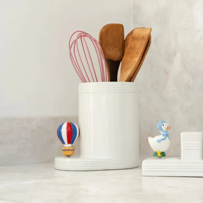 White container with wooden spoons and a whisk, small balloon figurines, and stacked white plates on a light background